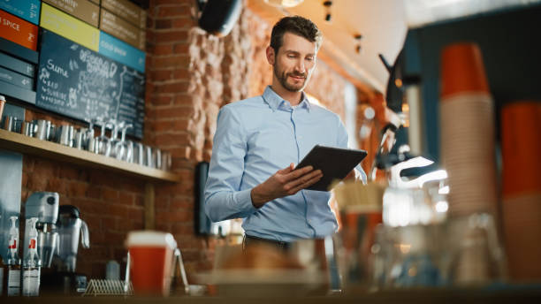 istock Handsome Caucasian Coffee Shop Owner is Working on Tablet Computer and Checking Inventory in a Cozy Loft-Style Cafe. Successful Restaurant Manager Standing Happy Behind Counter. 1293175106