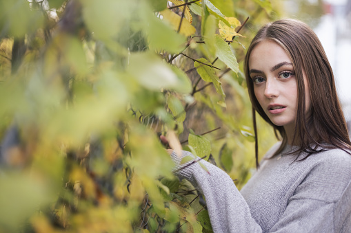 Attractive teenage woman standing at tall garden fence in between colorful autumn leaves looking over. Millennial Generation Female Autumn Portrait.