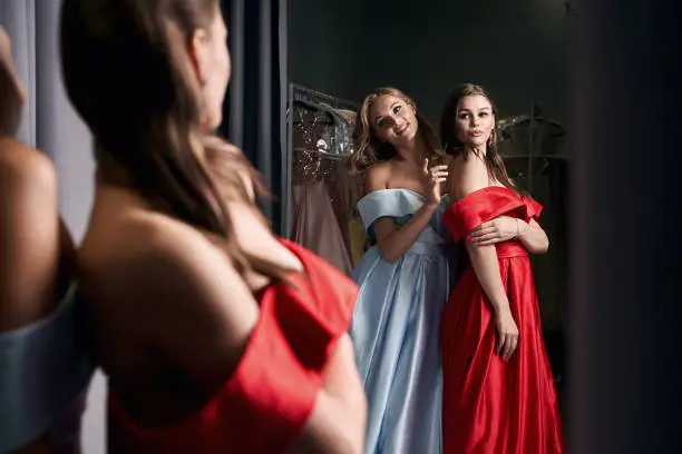 Two young beautiful women wearing off-the-shoulder full-length sky blue and crimson red satin slit prom ball gowns. Models looking in mirror.