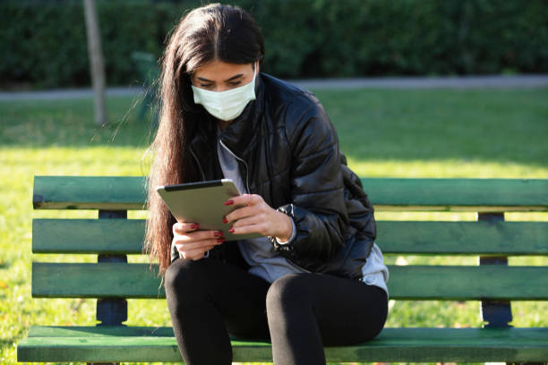 istock Young woman with face mask sitting on a bench in a public park using digital tablet 1293196721