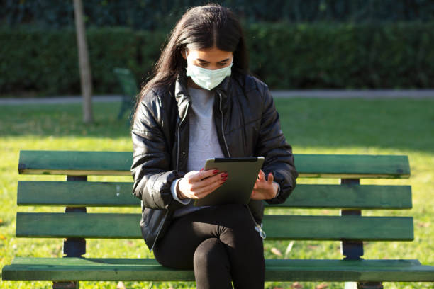 istock Young woman with face mask sitting on a bench in a public park using digital tablet 1293196872
