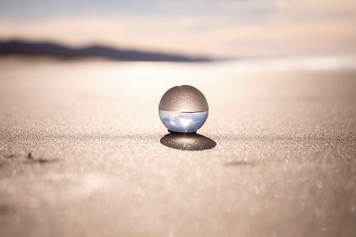 A glass ball in the sand is reflecting the water, sand, and sun behind it.