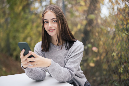 Happy smiling young woman sitting outdoors on white bench in autumn. Reading her social media post and e-mails on the smart phone. Looking up towards the camera with a happy smile. Millennial Generation Daily Social Media Lifestyle.