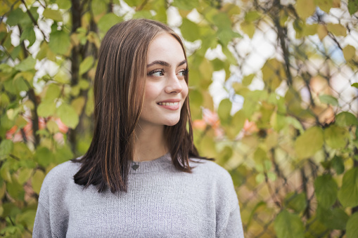 Bright happy smiling teenage woman standing outdoors in front of colorful autumn hedge looking over to her friends. Millennial Generation Female Autumn Portrait.