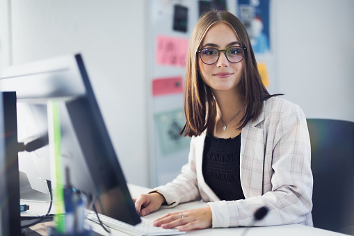 Businesswoman sitting in front of her computer monitor at her desk in home office looking up towards the camera. Young modern millennial gerneration businesswoman at work concept.