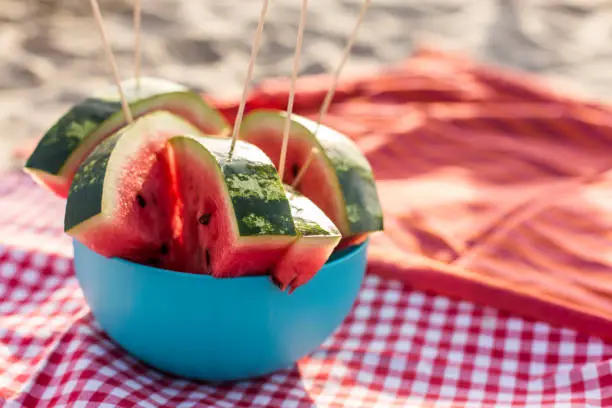 Copy space shot of delicious, juicy watermelon slices on a stick, arranged in a bowl, laying on a picnic blanket at the beach in warm summer sunshine.