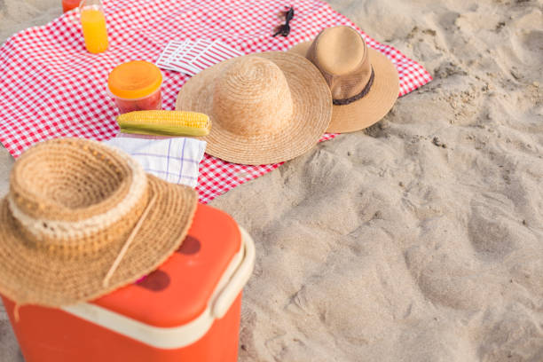 Flat lay of sun hats, sunglasses, corn on a cob, food cooler, cocktails and American flags arranged on a picnic blanket at the beach, ready to celebrate Independence day.
