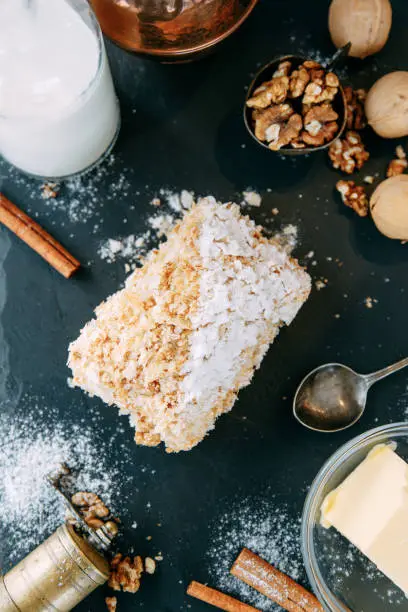 Photo of Napoleon cake with ingredients on a black stone Board. Napoleon cake with powdered sugar close-up.