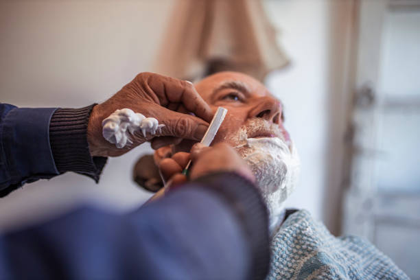 Skillful senior barber shaving beard by razor blade