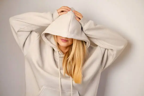 Photo of Young beautiful girl in a white hoodie posing. Warm oversized hoodie with an hood. Stylish trendy hipster bow. Trying on clothes in a store. Youth subculture. Fashion clothing advertising