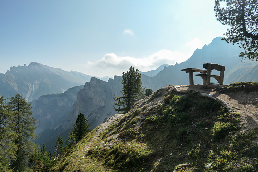 A vast mountain area in the Italian Dolomites. There is a wooden bench and a table at a small hill with the panoramic view on the Dolomites. Bright and sunny day. Few trees growing around it. Calmness