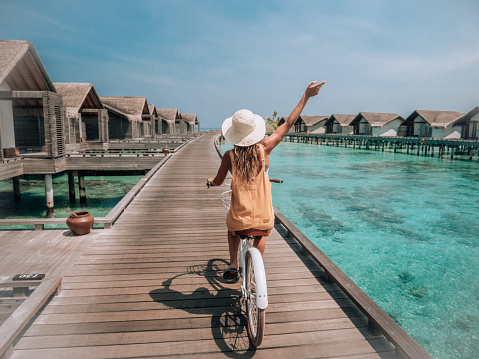 Tropical vacations young woman with bicycle on wooden pier in the Maldives contemplating overwater villas on the Island. Female enjoying nike ride on jetty over coral reef water. Dreamlike destination