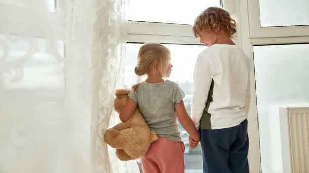 istock Two little kids in home clothes holding hands, looking out of a large window while spending time together at home 1293299876