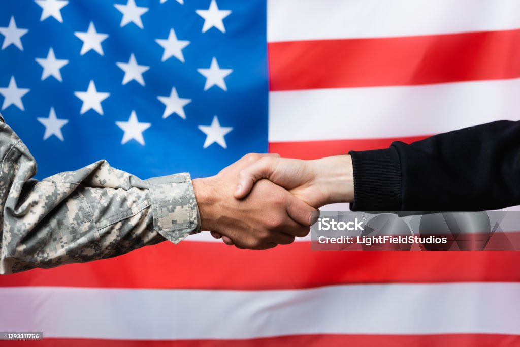 cropped view of soldier shaking hand with civilian man near american flag on blurred background Military Stock Photo