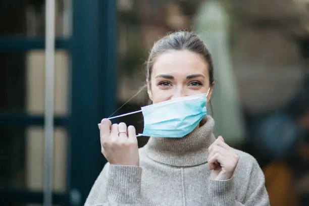 Portrait of a young attractive Caucasian woman standing outdoors and smiling while putting on a protective face mask