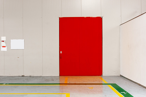 Empty industrial space with a large red metal door. The floor has green and yellow direction lines.