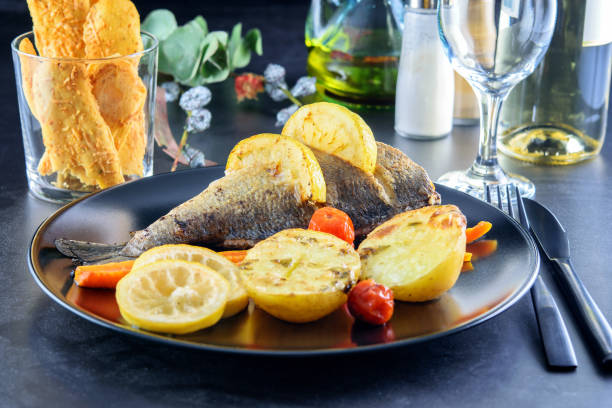 istock Black dish with fried fish dorada, slices of lemon, potatoes, carrot and little tomatoes cherry. Also there are cheese chips, black cutlery, wineglass, olive oil, a branch of eucalyptus on the dark table background. Festive decorated table. 1293329708