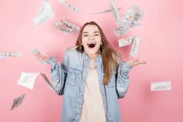 Photo of Portrait of pretty charming positive girl standing under shower from money having a lot of money in hands isolated on pink background