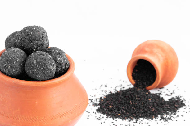Makar Sankranti Lohri Celebration - Kaala Til Gul Black Kala Laddu Decorated In Eco Friendly Mitti Terracotta Matka Or Terra Cotta Handi Pot And Whole Sesame Seeds Split On Floor On White Background