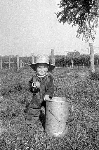 Baby boy dressed in overalls and hat standing next to milk pail outdoors on farm in summer 1927. Wellman, Iowa, USA.