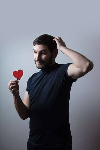 man in black shit holding red heart in tweezers with surprised and pensive face thinking about question, very confused idea