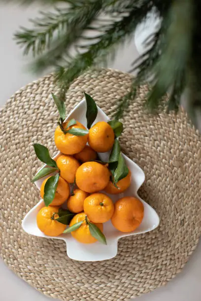 Photo of Small ripe tangerines with leaves, on plate in shape of Christmas tree on wicker serving napkin. Concept winter vitamin food. Moody still life festive composition. For greeting card, book, stories