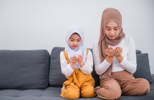Beautiful muslim young woman and cute girl sitting on couch and wearing hijab or headscarf with praying hands together in home. Mother and daughter prayer and praise for allah, muslim god.