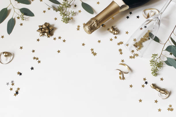 Happy New Year still life. Empty champagne glass, wine bottle with golden confetti stars and eucalyptus branches isolated on white table background. Celebration, party concept. Flat lay, top view. stock photo