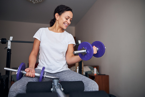 Joyful female bodybuilder working out with a pair of hand weights at her home gym