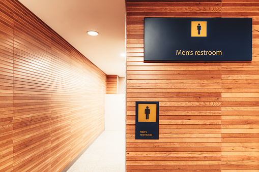 A bright interior with a wood cladding of a modern entrance to a public men toilet in an airport terminal, or a train station depot, or a mall, with a caption 