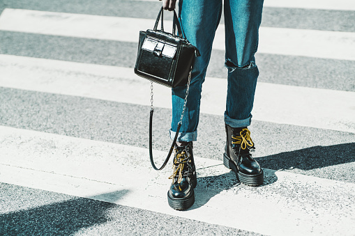 A close-up view of female legs in jeans and glossy boots with yellow laces - a woman standing on asphalt on the pedestrian zebra crosswalk with a small leather clutch in her hand on a sunny day