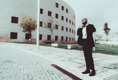 A fashionable virile bald black guy with a beard and in a custom formal suit is adjusting his blazer while standing on the paving-stone outdoors; with a bent building and copy space place on the left