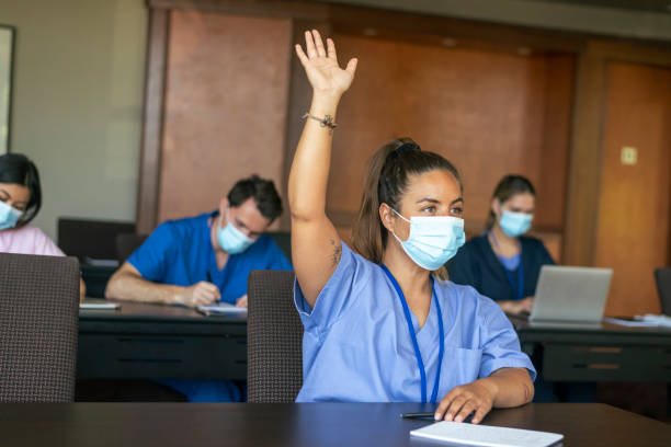 Female medical student raising hand in class A female medical student attending a school lecture raises her hand to ask a question. The multi-ethnic group of adult students is wearing medical scrubs and protective face masks to prevent viral infection during the Covid-19 pandemic. medical education stock pictures, royalty-free photos & images