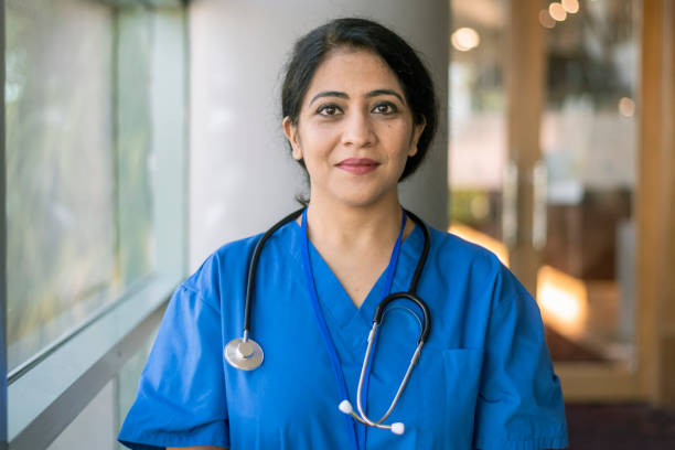 Portrait of confident ethnic female doctor A female doctor or nurse of Indian descent wearing scrubs and a stethoscope poses next to a window in a hospital corridor and smiles confidently directly at the camera. self sacrifice stock pictures, royalty-free photos & images