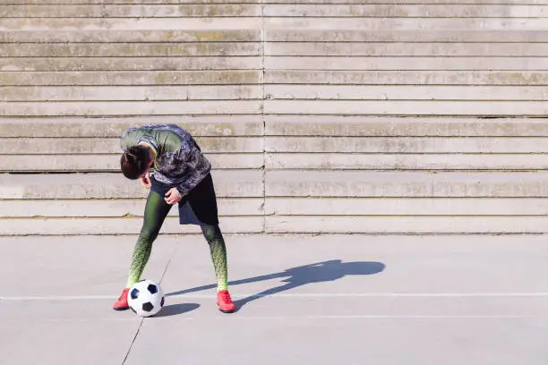 unrecognizable sportsman fitting his tights next to his football ball in a concrete soccer court, concept of healthy lifestyle and urban sport in the city, copy space for text