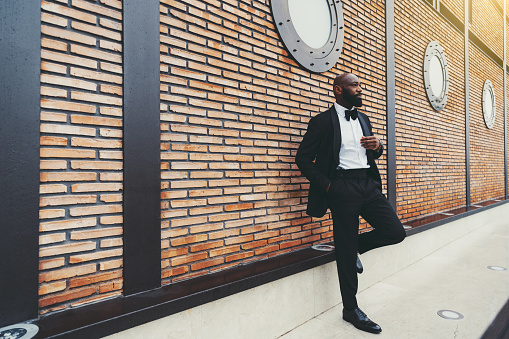 An elegant mature bald bearded African man in a black suit with a white shirt and a bow-tie is leaning against a modern brick wall with porthole windows above and with a copy space place on the left