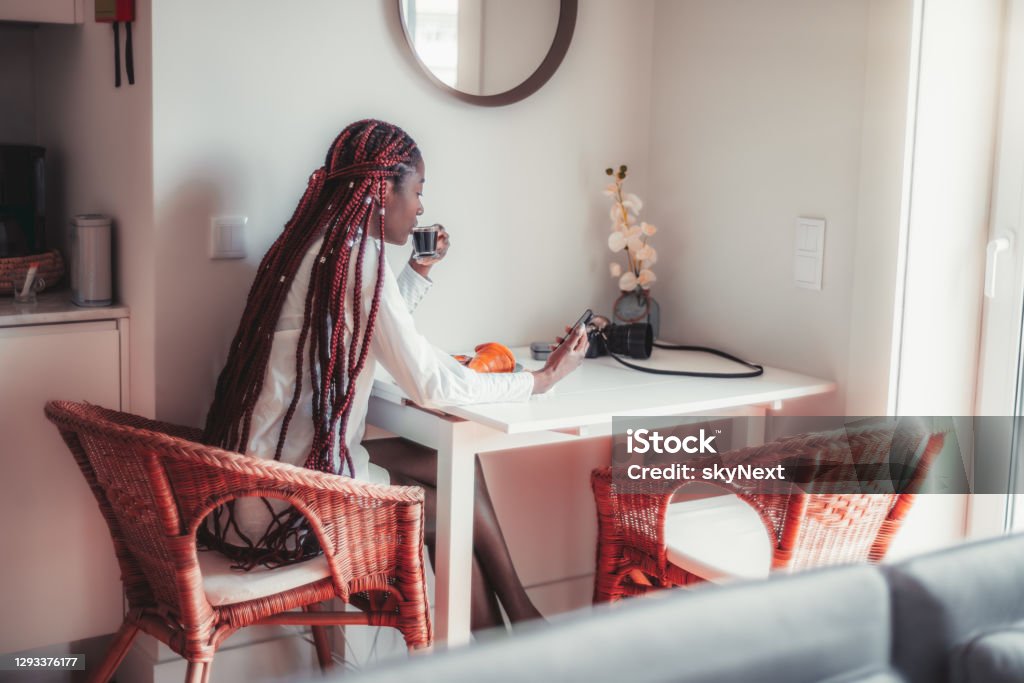 Black girl is having her breakfast A dazzling sensual young black female is having breakfast in the kitchen of her house, drinking an espresso and reading the news feed on the screen of her smartphone while sitting on wicker armchair Adult Stock Photo