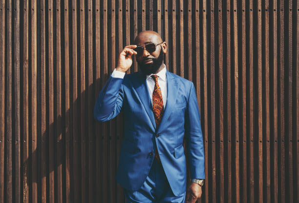 A black man in a fashion costume The portrait of a burly handsome bald black guy with a well-trimmed beard, in an elegant custom blue formal suit with a necktie, adjusting sunglasses outdoors in front of a wooden striped background necktie fashion adjusting suit stock pictures, royalty-free photos & images