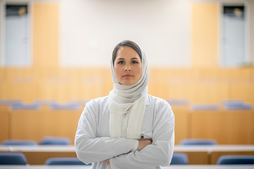 Young and confident female medical student posing in her lecture hall of her university. She wears a headscarf and a white lab coat.
