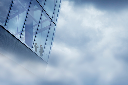A businesswoman and a businessman are seen through the windows of a tall office building that is enveloped by the clouds as they have a discussion.