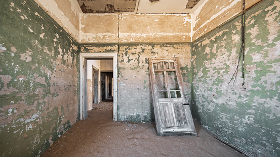 Abandoned old Diamond Mine Ghost Town Interior Panorama. Inside rotten abandoned old Diamond Mining Desert Building from the german empire years 1907-1928. Old wooden building door leaning on the run-down room. Nature is coming back. Desert Sand already entered the room and building. Kolmanskop, Luderitz, Namibia, Africa.