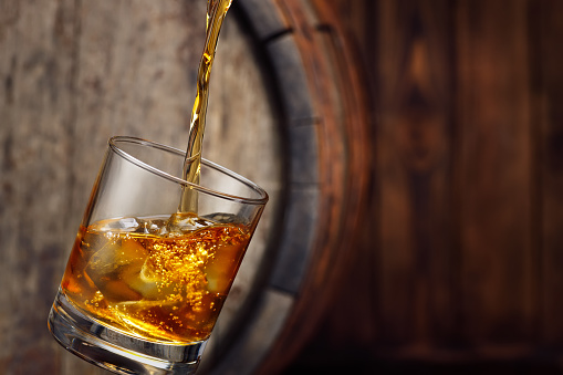 whiskey pouring into glass with ice and old wooden barrel on the background