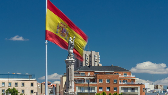 Spanish flag waves behind statue of Christopher Columbus timelapse with clouds on blue sky, plaza de Colon in Madrid, Spain