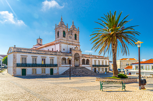 Sanctuary of Our Lady of Nazare catholic church in cobblestone square with palm trees in Sitio hilltop da Nazare town, clear blue sky in sunny summer day, Leiria District, Oeste region, Portugal