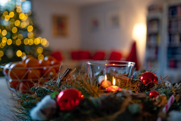 istock Cozy Christmas holidays home interior. Lit candle inside decorated wreath and clementines on table and sofa with red pillows and christmas tree at background. 1293441103