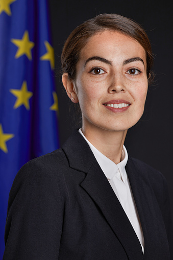 Vertical portrait of young female leader looking at camera and smiling while standing against EU flag in background