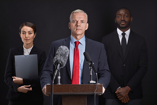 Waist up portrait of mature man giving speech standing at podium with two assistants in background