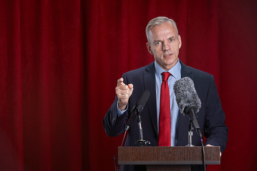 Waist up portrait of mature man speaking to microphone standing at podium on stage against red curtain, copy space