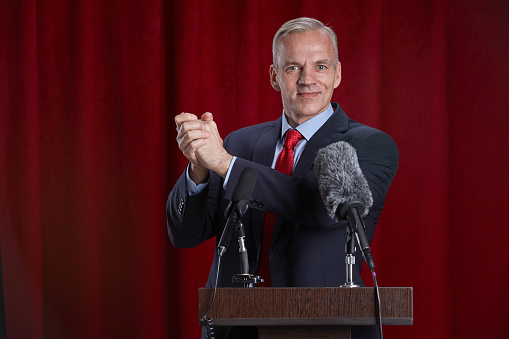 Waist up portrait of smiling mature man speaking to microphone standing at podium on stage against red curtain, copy space
