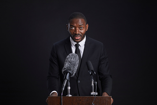 Waist up portrait of confident African-American man standing at podium and giving speech against black background, copy space